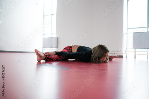 A girl stretching trainer lies in a transverse twine on a red gymnastic mat in a light fitness studio.