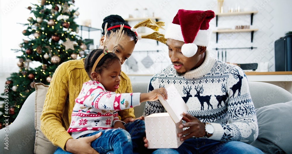 Portrait of curious African American kid girl opens gift box while sitting with happy parents together at decorated home near Christmas tree. New Year celebration, xmas present, holidays concept