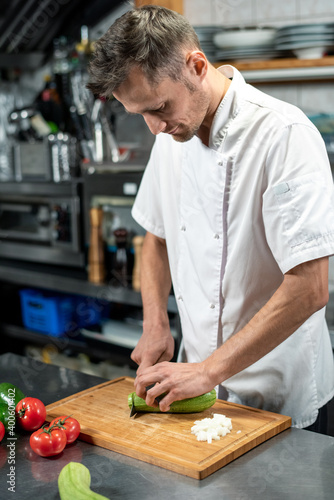 Young male chef in uniform bending over table while cutting fresh green zucchini