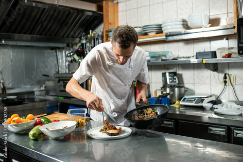 Young male chef with frying pan putting stew on plate with piece of fried salmon