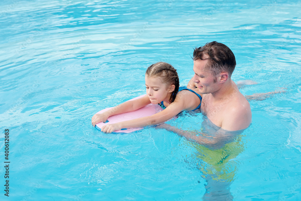 Father supporting little daughter with swimming board while teaching her to swim Father supporting little daughter with swimming board while teaching her to swim
