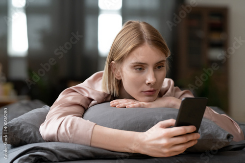 Young blond female in beige silk pajamas making selfie or scrolling through news