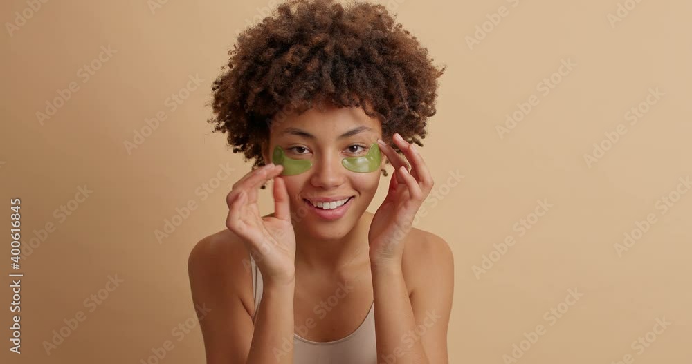 Beautiful young ethnic woman takes care of her skin takes off eye patches undergoes rejuvenation procedures poses against brown background wears collagen mask under lower eyelids. Beauty concept Beautiful young ethnic woman takes care of her skin takes off eye patches undergoes rejuvenation procedures poses against brown background wears collagen mask under lower eyelids. Beauty concept