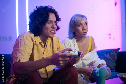 Young cheerful Caucasian husband and wife play videogames against each other at home enjoying leisure time