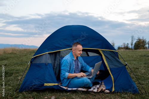 Man working on laptop in tent in nature. Young freelancer sitting in camp. Relaxing in mountains. Remote work, outdoor activity in summer. internet 5G.