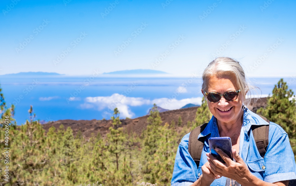 Happy active senior woman in mountain excursion with backpack sitting and using cellphone for a message. Horizon over sea and view of La Palma island