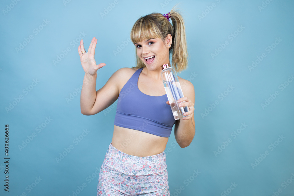 Young beautiful woman wearing sportswear holding a bottle of water over isolated blue background doing star trek freak symbol Young beautiful woman wearing sportswear holding a bottle of water over isolated blue background doing star trek freak symbol