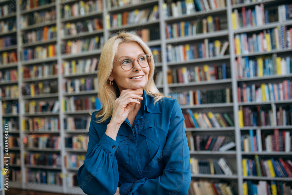 Portrait of beautiful female employee in eyeglasses and formal wear. Successful mature business woman stand in modern office looks away and smiling Portrait of beautiful female employee in eyeglasses and formal wear. Successful mature business woman stand in modern office looks away and smiling
