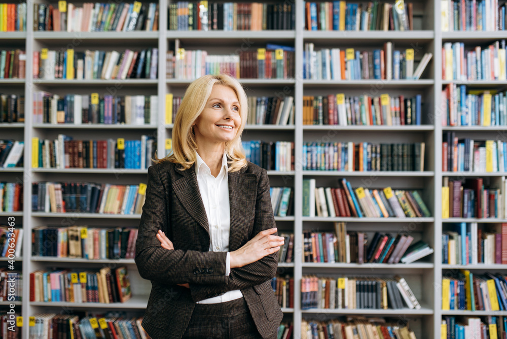 Beautiful mature lady with cross hands stands in modern office. Successful business woman in formal suit, employee or teacher looks away and smiling Beautiful mature lady with cross hands stands in modern office. Successful business woman in formal suit, employee or teacher looks away and smiling