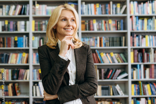 Close-up portrait of a gorgeous middle aged woman stand in modern office looks away and smiling. Beautiful female employee dressed in a formal suit thinking about new ideas for the work project