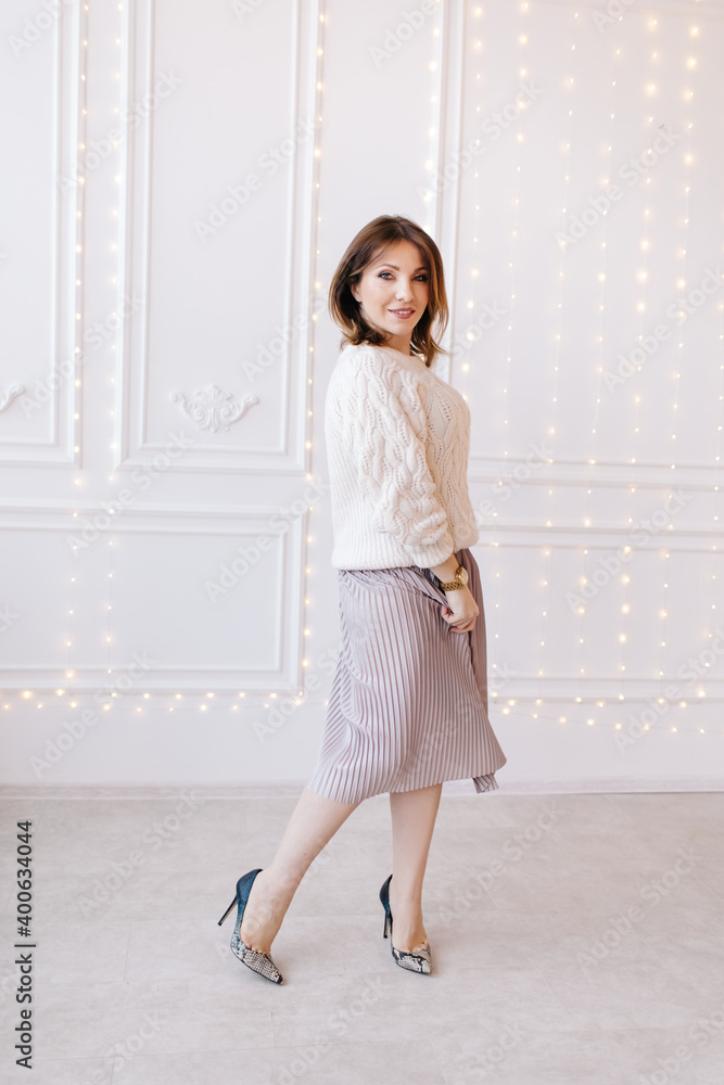 beautiful young woman in a white sweater, skirt and high heels on a background of white wall and christmas garlands. beautiful young woman in a white sweater, skirt and high heels on a background of white wall and christmas garlands.