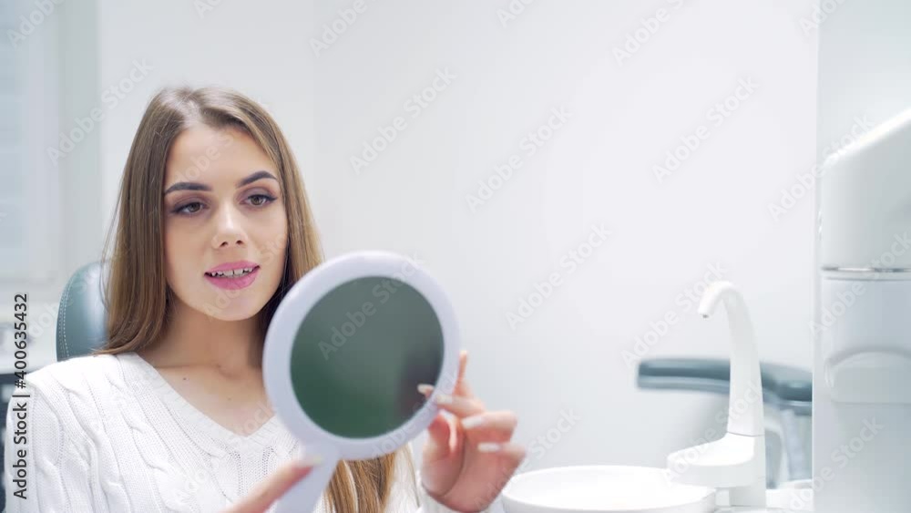 adult attractive girl patient in the dental office looks at the teeth in the mirror. Happy and smiling young woman examines teeth after whitening procedure or treatment while sitting on a chair adult attractive girl patient in the dental office looks at the teeth in the mirror. Happy and smiling young woman examines teeth after whitening procedure or treatment while sitting on a chair