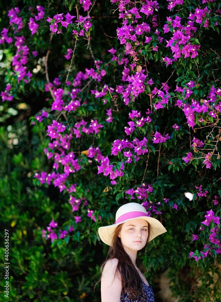 A young girl with long brown hair and a hat standing near to a blossimng tree.