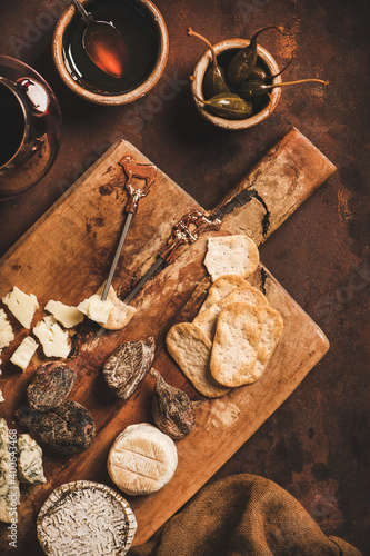 Assortment of snacks and appetizers for red wine concept. Flat-lay of various cheeses, crackers, smoked meat, fruit and glass of red wine on wooden rustic board over rusty brown background, top view