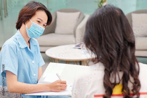 Positive medical nurse in protective mask talking to female patient and filling document