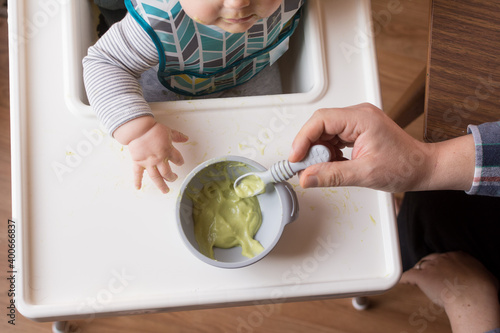 Baby sitting in high chair being offered first solid food avocado by Dad