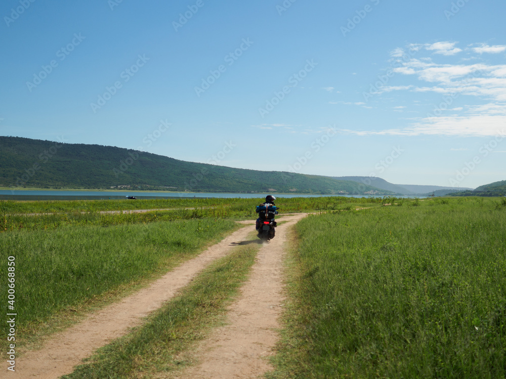 Fototapeta premium Young man rides a motorbike carrying equipment for a long vacation. In green meadows by lakes and valleys