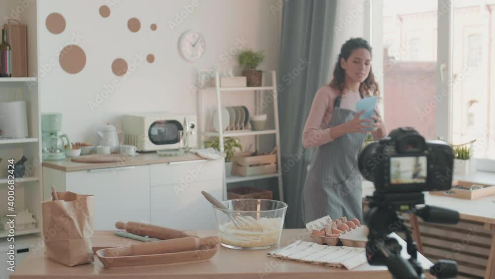 Medium long shot of brown-haired Caucasian female vlogger standing at kitchen table, walking to get water, adding it to flour in glass bowl, showing process on camera in front of her Medium long shot of brown-haired Caucasian female vlogger standing at kitchen table, walking to get water, adding it to flour in glass bowl, showing process on camera in front of her