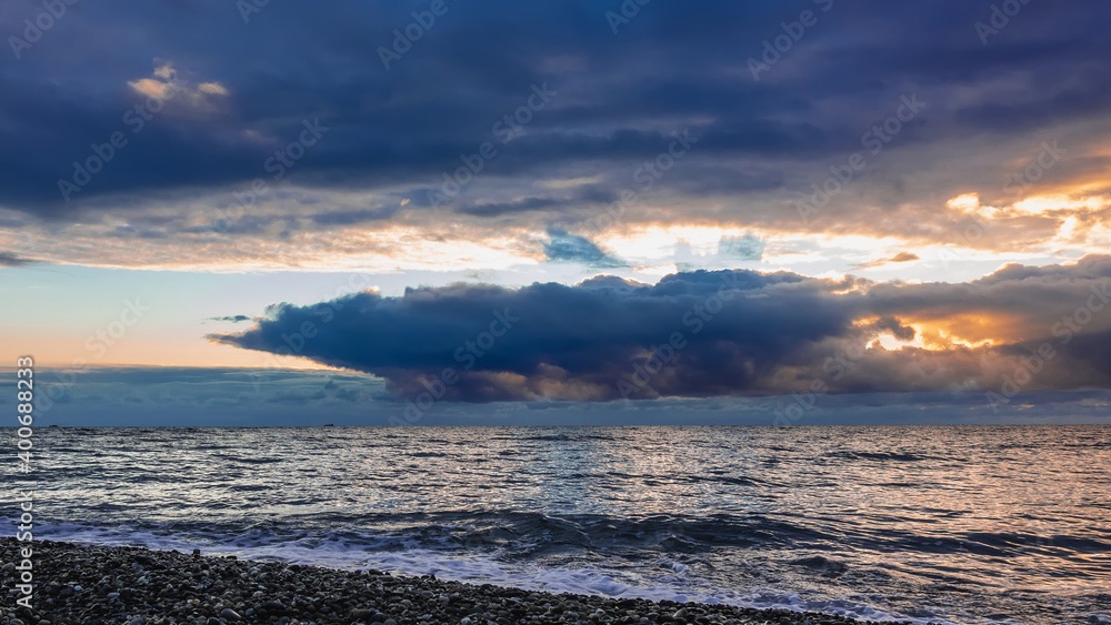Evening. The rays of the setting sun shine from behind dense blue clouds. The sky is backlit orange. Reflection on the shiny surface of the sea. Foam waves on the beach pebbles. Sunset. Black Sea. Evening. The rays of the setting sun shine from behind dense blue clouds. The sky is backlit orange. Reflection on the shiny surface of the sea. Foam waves on the beach pebbles. Sunset. Black Sea.
