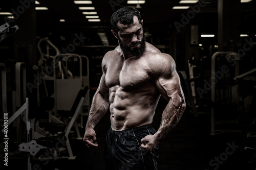 strong young bearded caucasian male with sport physique body standing in dark fitness gym with equipment