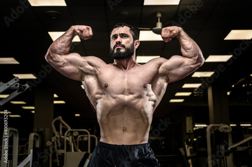 strong young caucasian man with beard showing powerful biceps arms muscle posing in dark fitness gym