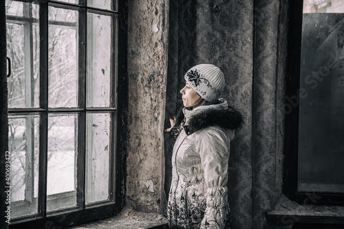 A girl stands at the window in an old abandoned building. Girl in a white jacket and hat. Old dirty window. The interior of an abandoned building.