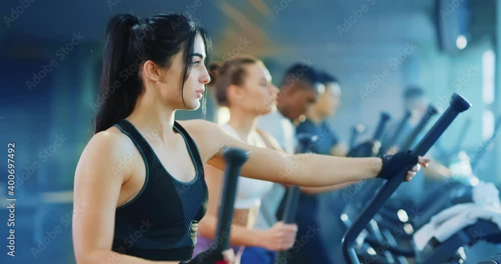 Team sports working out in modern gym. Portrait professional caucasian woman in sportswear training on cross trainer exercise bike cmiling into camera. Team sports working out in modern gym. Portrait professional caucasian woman in sportswear training on cross trainer exercise bike cmiling into camera.