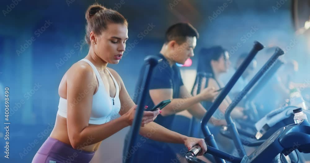 Young sportswoman trainin on eliptical bike using smartphone texting messages online application working out with other people in the gym. Young sportswoman trainin on eliptical bike using smartphone texting messages online application working out with other people in the gym.