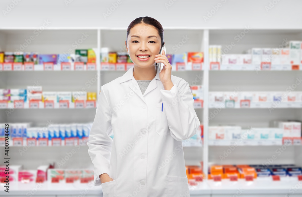 medicine, profession and healthcare concept - happy smiling asian female pharmacist or doctor calling on smartphone over pharmacy background medicine, profession and healthcare concept - happy smiling asian female pharmacist or doctor calling on smartphone over pharmacy background