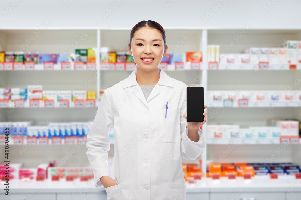medicine, profession and healthcare concept - happy smiling asian female pharmacist or doctor with stethoscope showing smartphone over pharmacy background