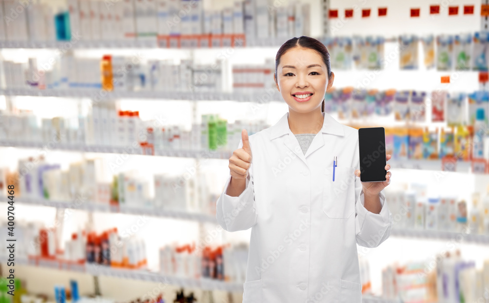 medicine, profession and healthcare concept - happy smiling asian female pharmacist or doctor with stethoscope showing smartphone and thumbs up over pharmacy background