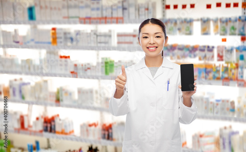 medicine, profession and healthcare concept - happy smiling asian female pharmacist or doctor with stethoscope showing smartphone and thumbs up over pharmacy background