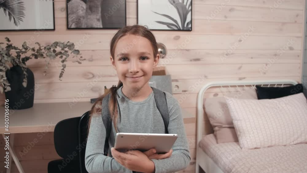 Portrait shot with handheld tracking of cute 8-year-old girl wearing backpack holding tablet and smiling for camera in her cozy room Portrait shot with handheld tracking of cute 8-year-old girl wearing backpack holding tablet and smiling for camera in her cozy room