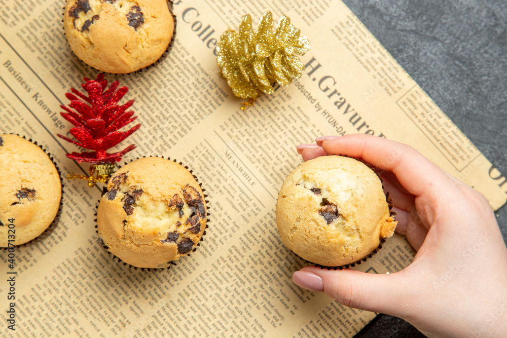 Hand holding one of delicious small cupcakes with accessories on newspaper on dark background