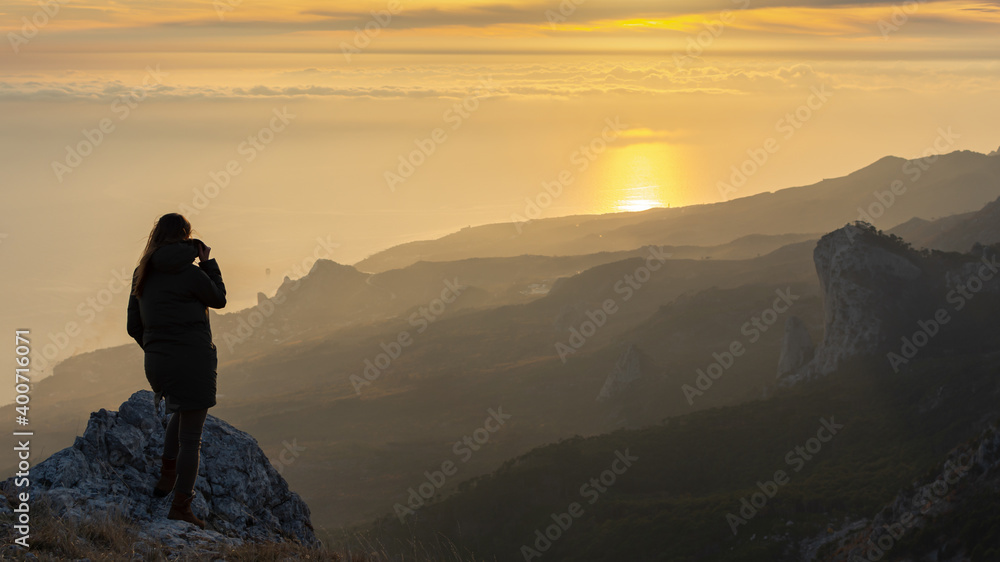 A girl stands on a mountain and looks at the sunset. Atmospheric evening view of the mountains and the sea. beautiful aerial perspective. The concept of relaxation and meditation. Bright sunlight.