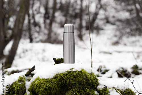 Steel vacuum thermos outside in the winter forest. Traveling aluminum flask standing on moss covered with snow.