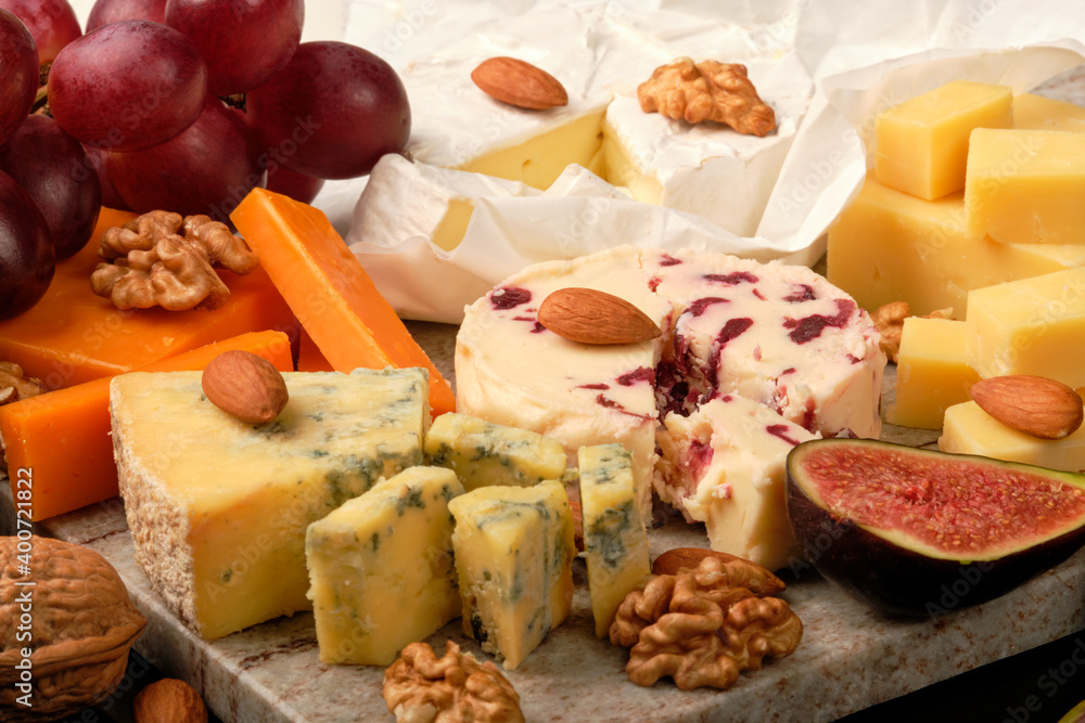 Cheese plate. Cheddar, Brie, Red Leicester, Wensleydale with sweetened dried cranberries, Blue Stilton, almond and walnut on a marble board with grape and figs on a black background.