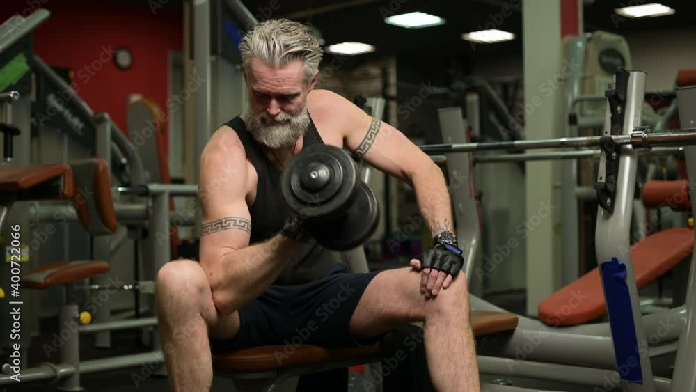 A gray-haired man with a beard and tattoos pumps biceps in the gym using a dumbbell A gray-haired man with a beard and tattoos pumps biceps in the gym using a dumbbell