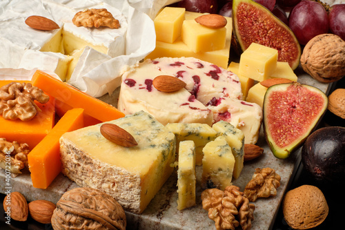 Cheese plate. Cheddar, Brie, Red Leicester, Wensleydale with sweetened dried cranberries, Blue Stilton, almond and walnut on a marble board with grape and figs on a black background.