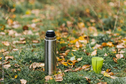 A metal thermos with hot tea and a green plastic cup stand on the grass with yellow maple leaves against the backdrop of nature. Autumn portrait, advertising.