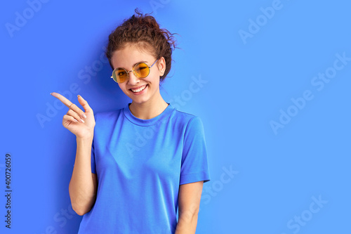 positive smiling teen girl pointing fingers at side, index at left. female in sunglasses isolated over blue background