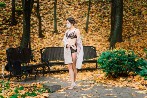 Young beautiful dark-haired woman with slender figure in lingerie and classic coat in autumn park