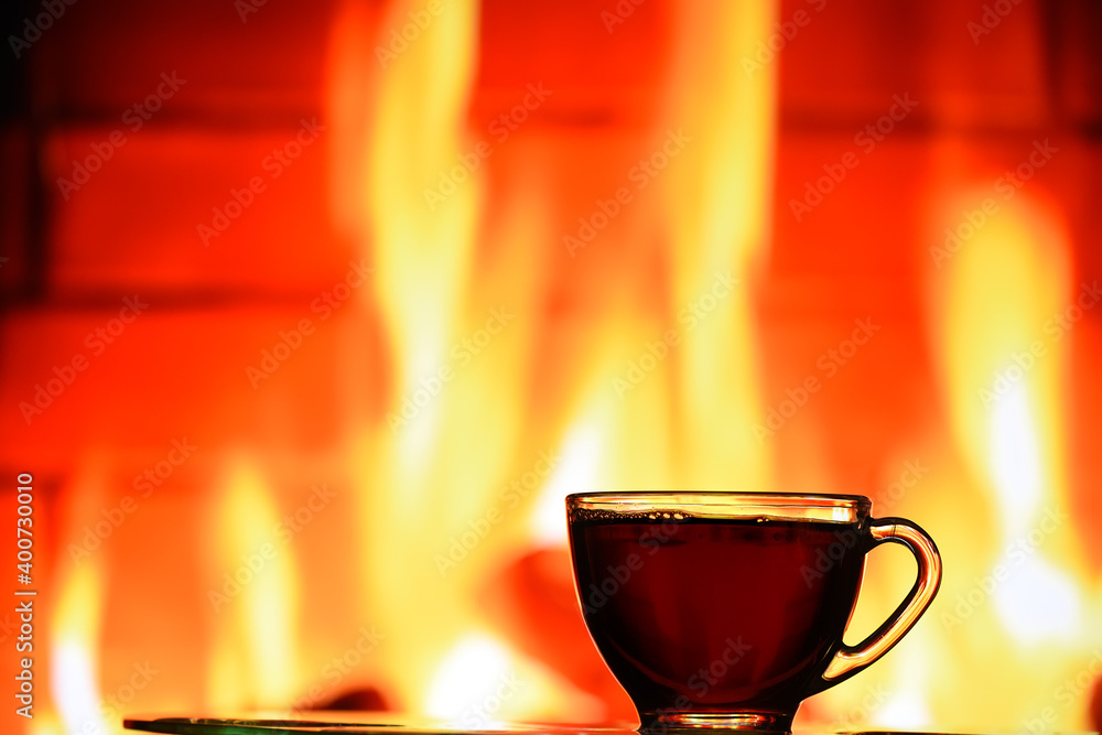 A transparent cup with black tea against the background of a burning fireplace. Evening winter photo by the fireplace. The atmosphere of a cozy winter evening at home.