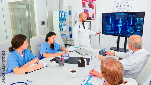 Mature medical physician explaining treatment to nurses during healthcare seminar pointing at digital monitor. Clinic herapist discussing with colleagues about disease, medicine professional.