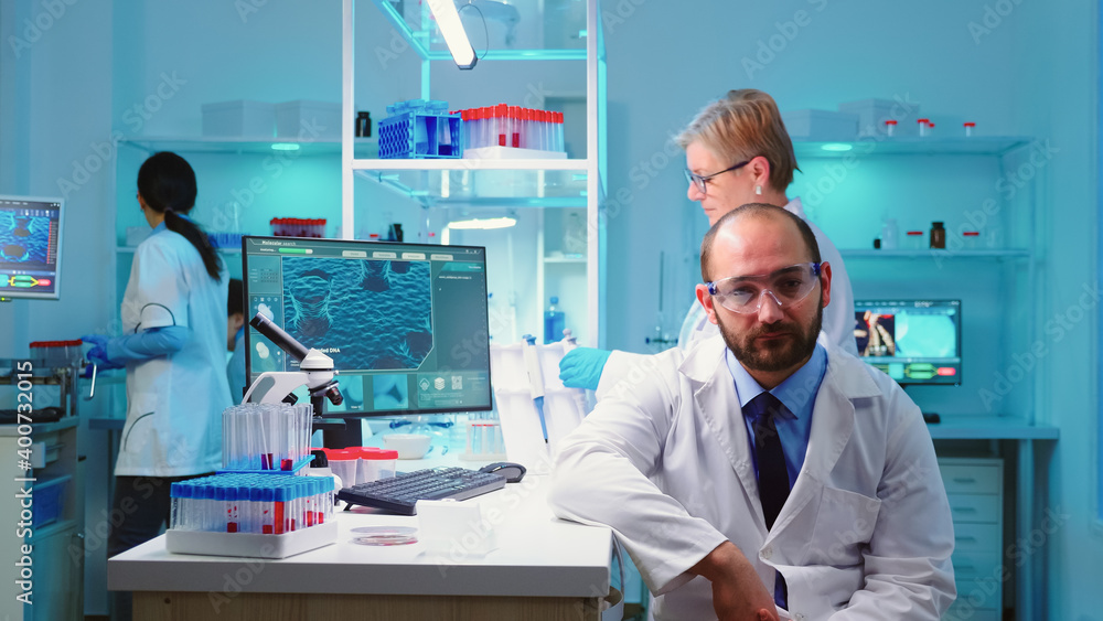 Portrait of experienced scientist man looking tired at camera working overtime in equipped lab. Examining virus evolution using high tech and chemistry tools for vaccine development.