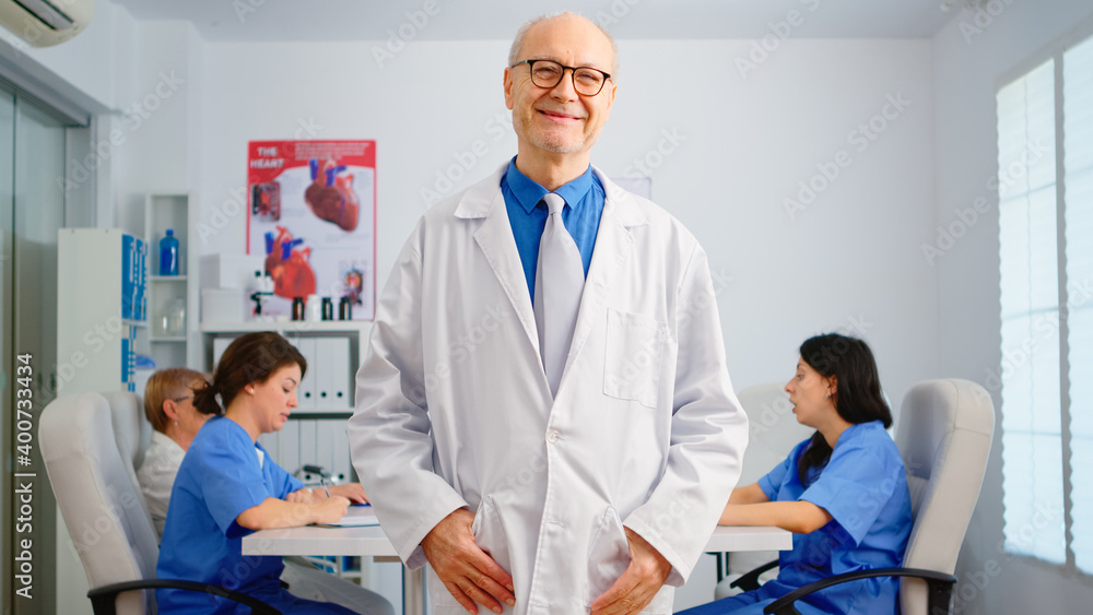 Obraz premium Portrait of elderly man surgeon standing in front of camera smiling in medical conference meeting office. Team of doctors working in background talking about symptoms of disease in clinic room