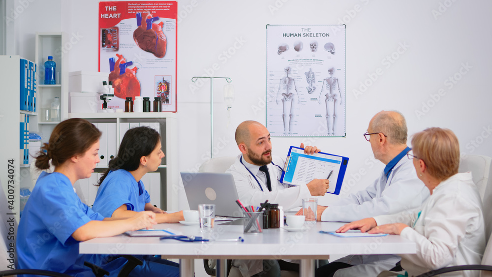 Doctor is brainstorming to analyze medical problems pointing on clipboard showing to colleagues medical statistics. Coworkers working in hospital office discussing symptoms of disease, taking notes. Doctor is brainstorming to analyze medical problems pointing on clipboard showing to colleagues medical statistics. Coworkers working in hospital office discussing symptoms of disease, taking notes.