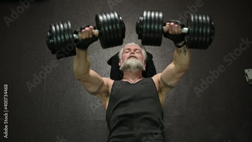An overhead view of an athletic gray-haired man with a beard and tattoos, pressing dumbbells on a bench, training his chest muscles in the gym