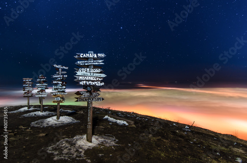 Arrows with direction indicators on the top of Mashuk mountain at night. Low clouds and fog are painted with bright colors from the light of city lanterns. A meteor is visible. Long exposure. Russia