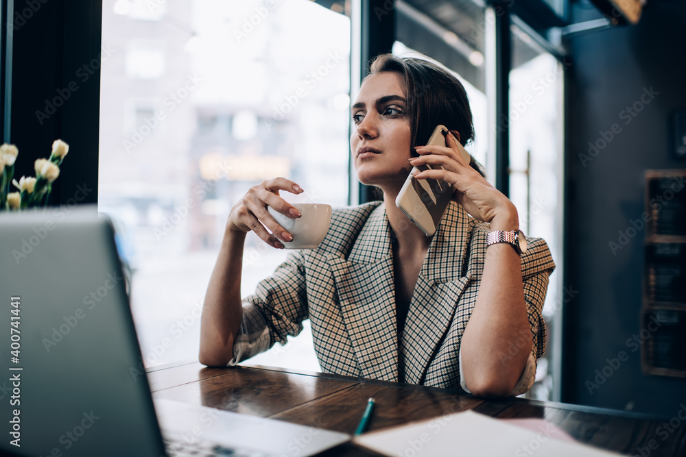 Smart casual manager with coffee cup making business conversation via smartphone application for discussing delivery order, Caucasian female small owner sitting at cafe table with laptop and calling Smart casual manager with coffee cup making business conversation via smartphone application for discussing delivery order, Caucasian female small owner sitting at cafe table with laptop and calling